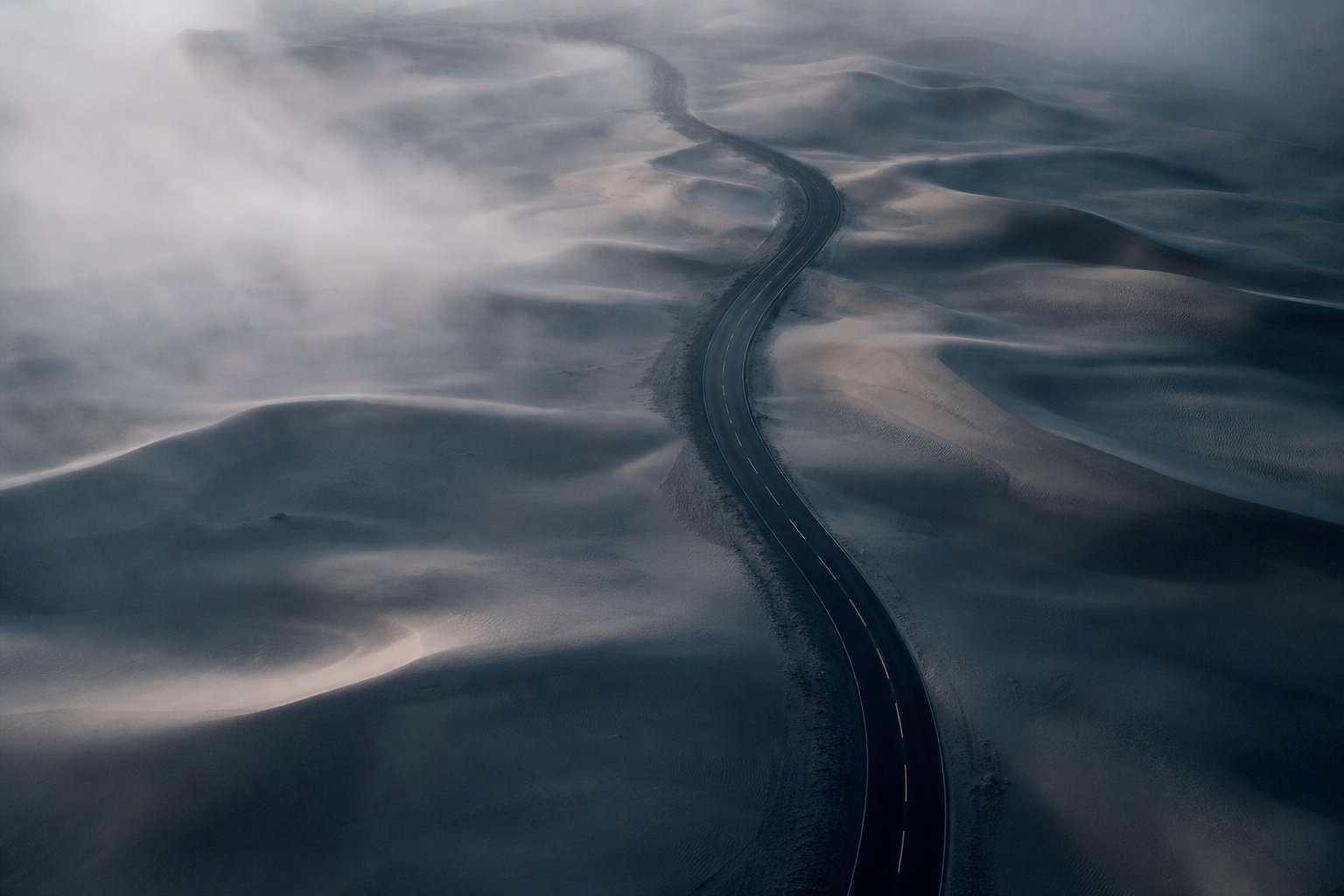 Aerial view of a winding road through cool, foggy sand dunes at dawn